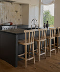 A modern kitchen features a dark island with a sink, Hampton Counter Stools along the counter, a light botanical-patterned tiled backsplash, and a large window framing views of lush greenery.