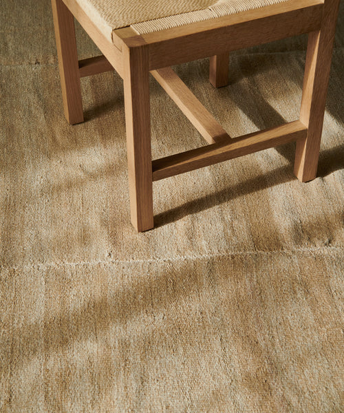 A close-up of a wooden chair with a woven seat rests on the Handwoven Vintage Rug, its intricate texture highlighted by sunlight and gentle shadows on the floor.