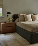 A modern bedroom features a neatly made bed with beige linens, a dark frame, a wooden nightstand with vases of white flowers, a bowl, book, black wall lamp, and is anchored by the Handwoven Vintage Rug.