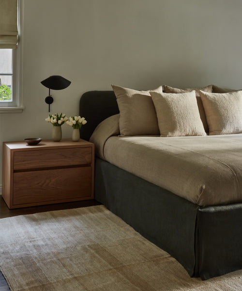 A modern bedroom features a neatly made bed with beige linens, a dark frame, a wooden nightstand with vases of white flowers, a bowl, book, black wall lamp, and is anchored by the Handwoven Vintage Rug.