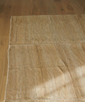 The Handwoven Vintage Rug in beige, featuring visible seams, rests on a light brown wooden floor while natural sunlight casts soft shadows across the surface.