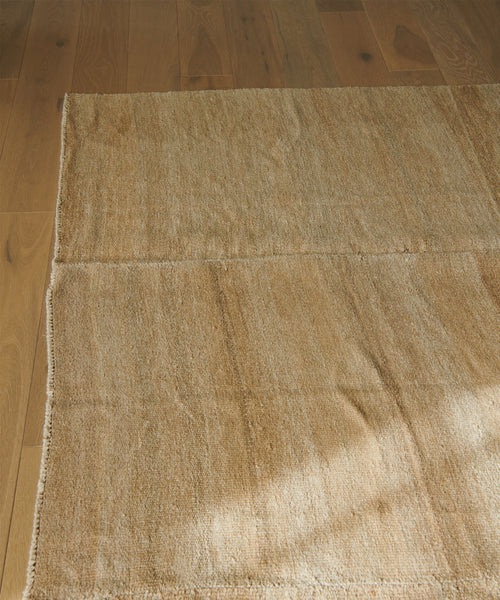 The Handwoven Vintage Rug in beige, featuring visible seams, rests on a light brown wooden floor while natural sunlight casts soft shadows across the surface.