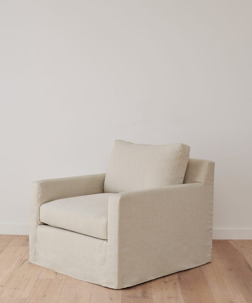 The Harbor Chair Slipcover in light beige natural linen covers a modern upholstered armchair with cushioned seat and backrest, shown on a wooden floor against a plain white wall.