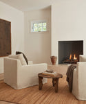 A cozy living room features two Harbor Chair Slipcovers in natural linen facing a modern fireplace, with a rustic wooden coffee table, books, a brown vase, a textured rug, and a small window on the cream wall.
