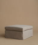 A square upholstered ottoman or stool cushion in a light taupe-beige fabric, shown against a solid warm beige studio background.
