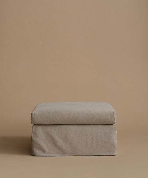 A square upholstered ottoman or stool cushion in a light taupe-beige fabric, shown against a solid warm beige studio background.