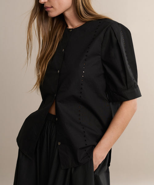 A woman with long hair wears the black Harper Blouse in Italian cotton poplin with button details and a matching skirt, standing against a beige background. Her face is partially out of frame.