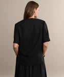 A woman with long brown hair wears a loose black Harper Blouse and a matching skirt, standing with her back to the camera against a plain beige background.