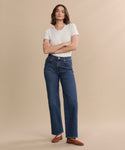 A person stands against a beige background wearing a white short-sleeve t-shirt, the Harper Jean in organic cotton with a wide-leg silhouette, and brown shoes, arms crossed and looking at the camera.