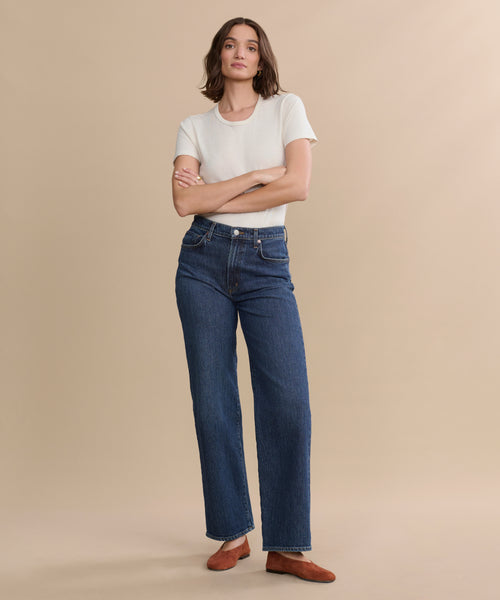 A person stands against a beige background wearing a white short-sleeve t-shirt, the Harper Jean in organic cotton with a wide-leg silhouette, and brown shoes, arms crossed and looking at the camera.