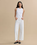 A woman with long dark hair stands against a beige background, wearing the high-waisted Harper Jean in organic cotton with a wide-leg silhouette, a white sleeveless top, and black flats. Her hands are in her pockets.