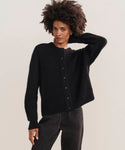A woman with short curly hair stands against a plain background wearing the Hayes Crewneck Cardigan in black and dark jeans, calmly gazing at the camera with one hand resting behind her head.