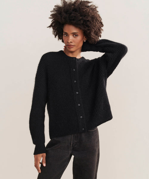A woman with short curly hair stands against a plain background wearing the Hayes Crewneck Cardigan in black and dark jeans, calmly gazing at the camera with one hand resting behind her head.