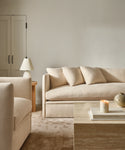 A minimalist living room with the Dume Sofa in beige sustainable hemp, a matching armchair, light wood side table with lamp, and a travertine coffee table displaying books, a candle, and a neutral-toned vase.