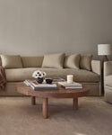 A neutral-toned living room with the Dume Sofa in sustainable upholstery, a round wooden coffee table with books, a lit candle, white flowers in a vase, and a bowl. A lamp is on the side table in the background.