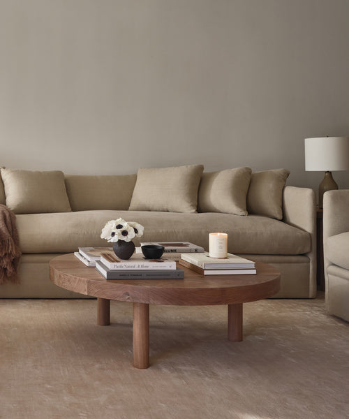 A neutral-toned living room with the Dume Sofa in sustainable upholstery, a round wooden coffee table with books, a lit candle, white flowers in a vase, and a bowl. A lamp is on the side table in the background.