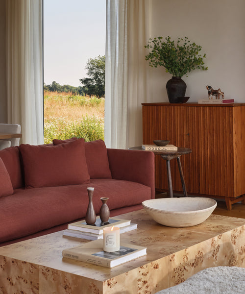 A cozy living room with a rust-colored sofa, light wood coffee table featuring the Hudson Glass Candle, a wooden cabinet, a vase with greenery, and large window curtains revealing sunlight over the grassy field outside.