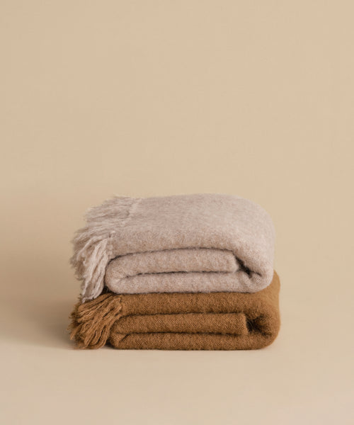 Two wool throws—a lightweight light beige alpaca throw and a brown Hudson Throw—are stacked atop each other against a plain beige background. Both blankets have fringed edges.