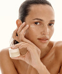 A woman with smooth skin holds a small jar of Hydra Eye Balm near her face, gently touching her cheek. With slicked-back hair and minimal makeup, she demonstrates how to reduce puffiness and dark circles against a light background.