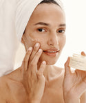 A woman with a towel wrapped around her head applies cream to her cheek while holding a jar labeled The Hydration Heroes, looking at the camera and smiling softly.