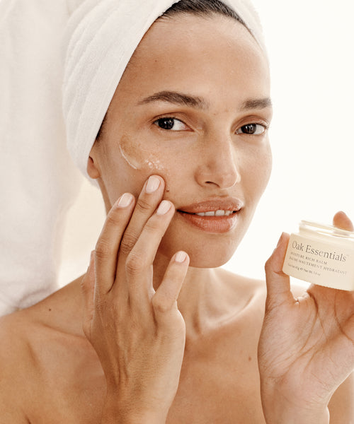 A woman with a towel wrapped around her head applies cream to her cheek while holding a jar labeled The Hydration Heroes, looking at the camera and smiling softly.