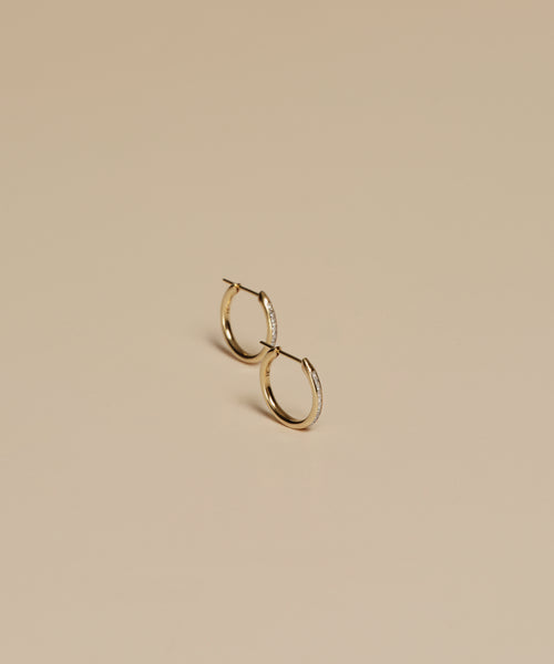 The Iris Diamond Hoops are small 14k gold earrings with subtle embedded stones, elegantly displayed on a smooth beige surface.