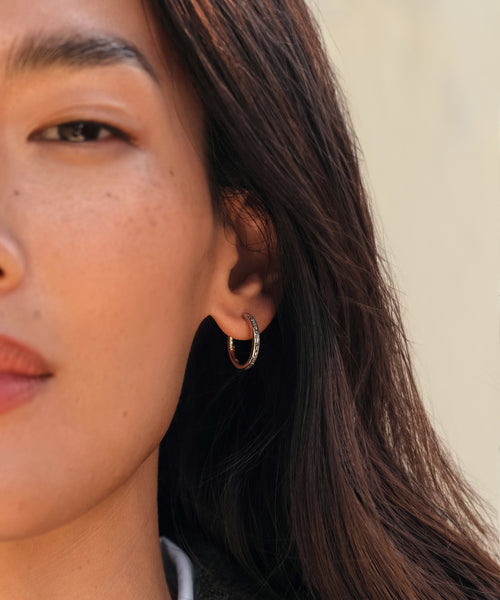 Close-up of a person with long brown hair wearing the Iris Diamond Hoops by Jenni Kayne. The focus is on one side of their face, highlighting the gold hoops adorned with diamonds and their clear skin.