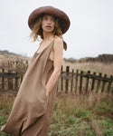 Woman in a brown dress and hat standing in a field with a wooden fence in the background
