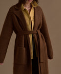 A woman wears the Jessica Sweater Coat, a brown alpaca blend long coat, over a yellow button-up shirt, standing against a plain beige background. Her face is partially out of frame.