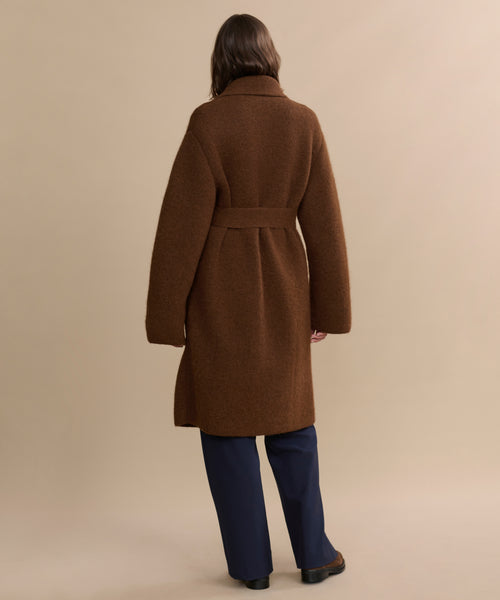A person with long brown hair stands with their back to the camera against a beige background, wearing navy pants and the brown, belted Jessica Sweater Coat made from an alpaca blend.