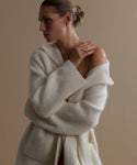 A woman with short, slicked-back hair models the Jessica Sweater Coat in ivory, paired with minimal gold jewelry. She stands against a neutral background with arms crossed, gazing thoughtfully to the side.