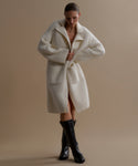 Against a beige backdrop, a person wears the Jessica Sweater Coat—an oversized, long ivory alpaca-blend piece—paired with black knee-high boots. Their head is slightly bowed as they adjust the front of the coat.