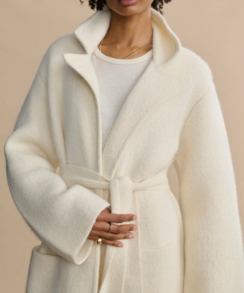Wearing the Jessica Sweater Coat in ivory over a white top, accessorized with gold rings and a delicate gold necklace, a person poses with one hand resting on the belt against a neutral background.