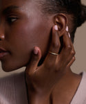 A woman in profile touches her ear, showing smooth dark skin, natural makeup, pale pink nails, and a June Baguette Band on her finger. She wears a light knit sweater.