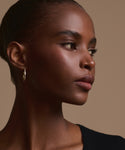 A woman with smooth dark skin and braided hair looks to the side, wearing the June Baguette Hoops in 14k gold and a black top, against a plain light tan background.