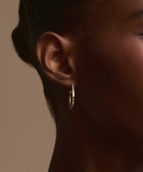 Close-up of a person's ear wearing June Baguette Hoops—14k gold earrings with embedded gemstones, inspired by baguette diamond styles. The neutral background frames part of the face, ear, and neck.