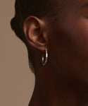 Close-up of a person's ear wearing Jenni Kayne's June Baguette Hoops—a small silver hoop earring—against smooth dark skin and pulled-back hair, set against a plain beige background.