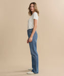 A woman in profile stands against a beige background, wearing a fitted white tee and AGOLDE Kelly Jean high-rise straight jeans. She has shoulder-length wavy hair, white shoes, and a neutral expression.