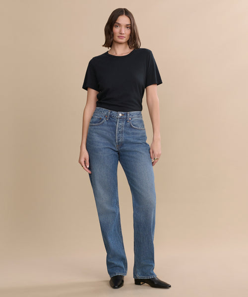 A woman with shoulder-length brown hair stands against a beige background wearing a plain black t-shirt, black boots, and the sustainable denim AGOLDE Kelly Jean high-rise straight jeans, posing with a relaxed posture.