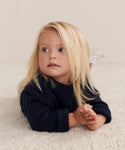 A young child with long blonde hair lies on a soft, light-colored carpet, wearing the cozy Kids Cashmere Fisherman sweater in navy and gazing upward with a thoughtful look.