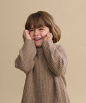 A young child with brown hair smiles joyfully, hands on their cheeks, while wearing a cozy Kids' Cashmere Fisherman sweater against a plain beige background.