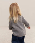 A young child with long blonde hair, wearing a heather grey Kids Cashmere Fisherman sweater and dark blue pants, stands with their back to the camera on a light-colored carpet.