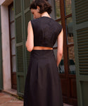 A woman with short dark hair stands outside by green shutters, showing off the Kit Dress—a sleeveless black summer staple. She faces away to highlight the chic back details of this must-have ensemble.