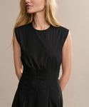 A woman with long blonde hair models the sleeveless black Kit Dress against a beige background, looking slightly to the side with her hands behind her back, showcasing this timeless summer staple.
