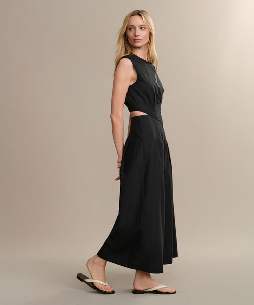 A woman in a sleeveless black Kit Dress with side cutouts poses in profile against a beige background, wearing white flip-flops and glancing over her shoulder.
