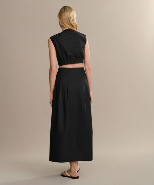 A person with long blonde hair stands with their back to the camera, wearing a sleeveless black Kit Dress—a perfect summer staple—paired with black sandals against a plain beige background.