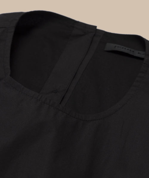 Close-up of the Kit Dress: a black dress with a round neckline and visible collar seam, laid flat on a neutral beige background. A small black label is visible near the neckline—an effortless summer staple.
