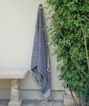 The Laguna Beach Towel, featuring black and white stripes and fringed edges, hangs on a light wall near a green leafy bush and a stone bench.