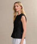 A woman with long blonde hair models the sleeveless black Lara Top and white pants, standing sideways against a plain beige background, gazing at the camera with a neutral expression.