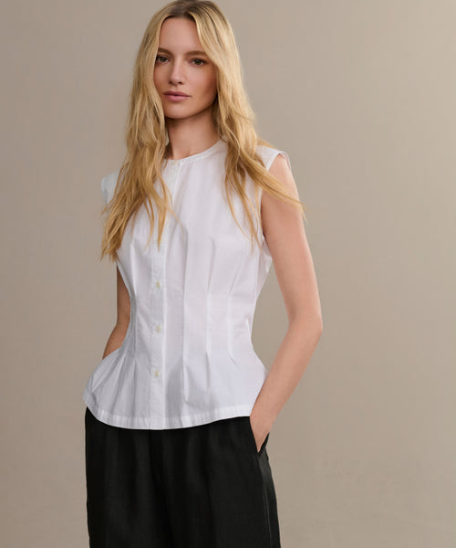 A woman with long blonde hair wears the Lara Top, a sleeveless button-front blouse, paired with black pants and standing with one hand in her pocket against a plain beige background.